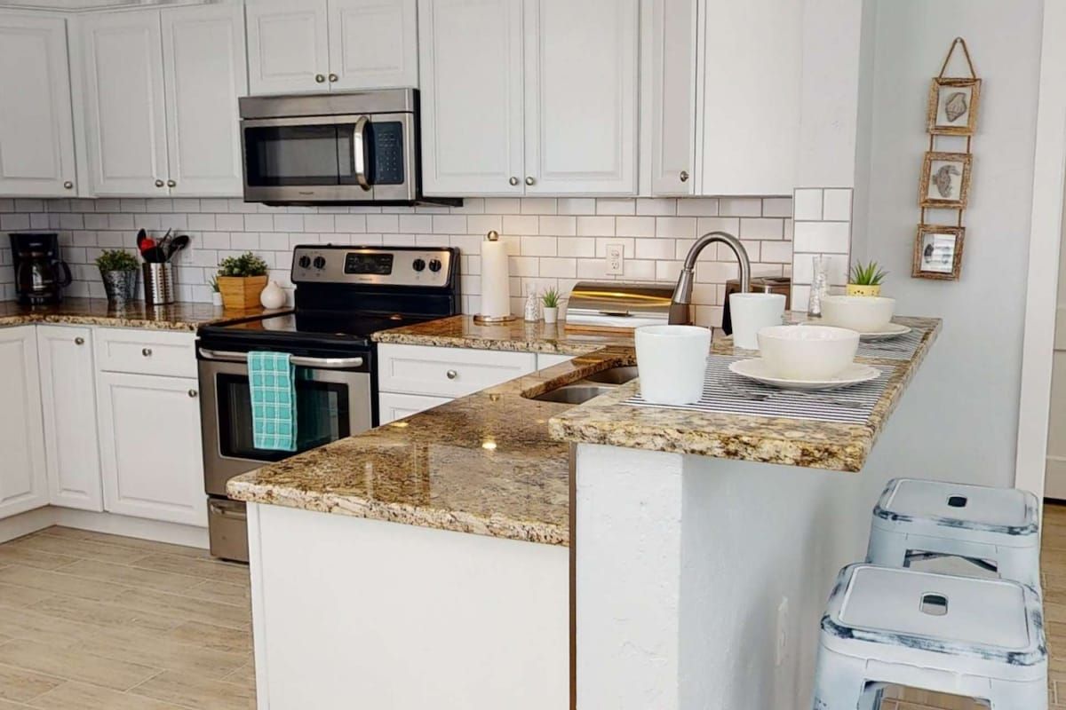 A kitchen with white cabinets, a stainless steel microwave, a black stove, granite countertops, and a small island with stools.