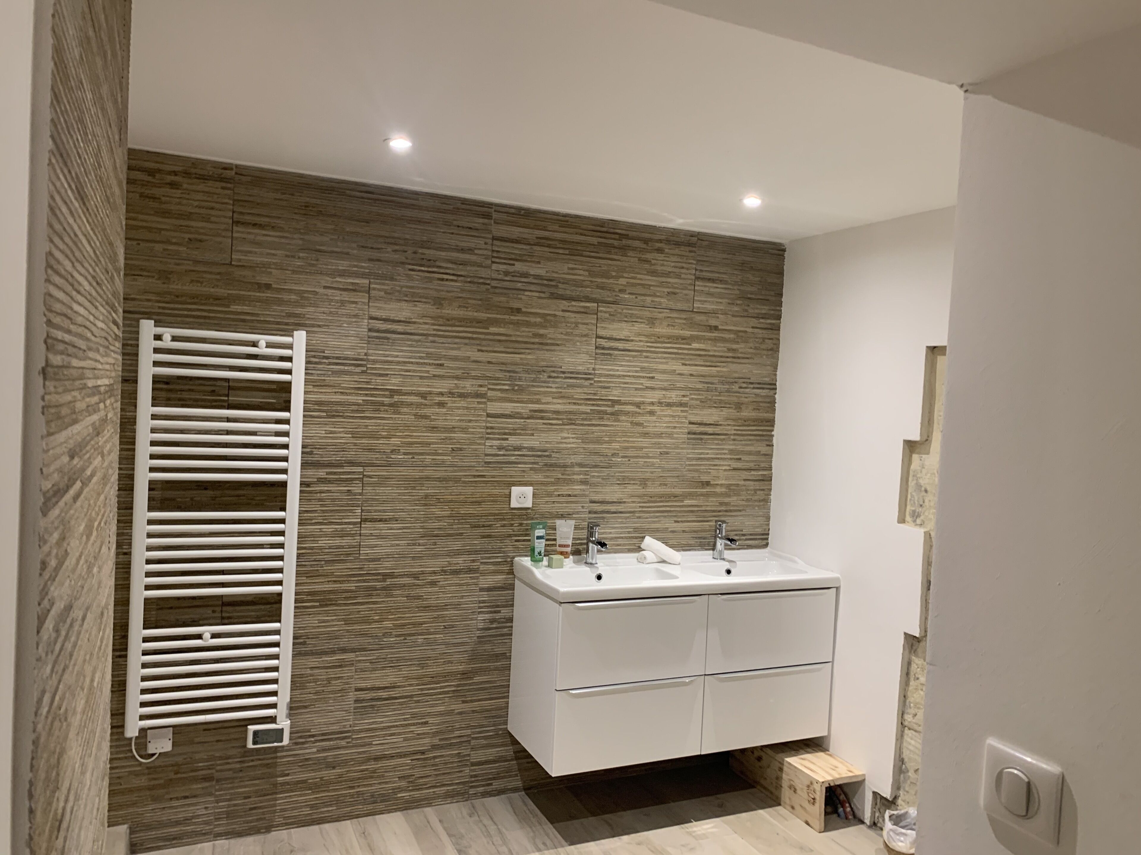 A modern bathroom with a wooden wall, a white sink with two basins, and a towel rack.