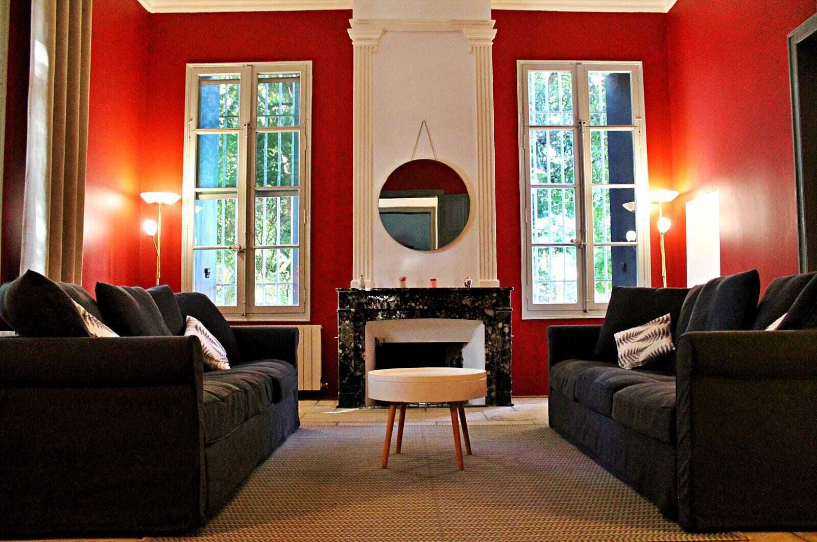 A room with two sofas facing a fireplace, large windows with curtains, and a round mirror above the fireplace.