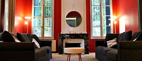 A room with two sofas facing a fireplace, large windows with curtains, and a round mirror above the fireplace.