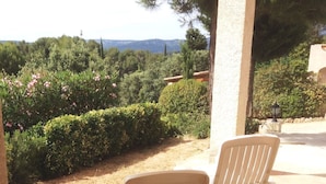 Deux fauteuils de jardin blancs sont disposés sur une terrasse carrelée, offrant une vue sur un jardin et des collines lointaines.
