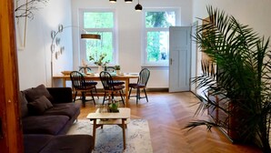 A living room with a sofa, a coffee table, and a dining area with chairs and a table. There is a large plant in the corner and a window with a view of trees.