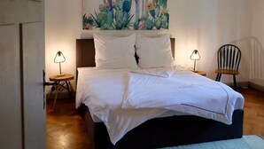 A bedroom with a bed, two bedside tables, a chair, a wall art of cacti, and a hanging light fixture.