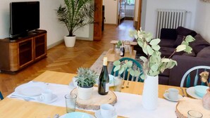 A dining area with a wooden table set for four, surrounded by chairs. There is a television on a stand to the left and a potted plant in the center.