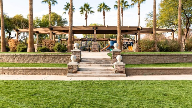 A covered outdoor area with steps leading to a playground, surrounded by palm trees and greenery. - Cute, Clean & Comfortable in the Best Community! (Gilbert)