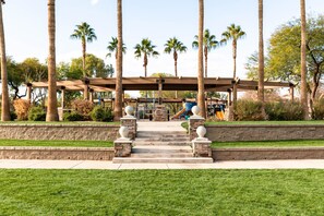 A covered outdoor area with steps leading to a playground, surrounded by palm trees and greenery. - Cute, Clean & Comfortable in the Best Community! (Gilbert)