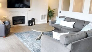Uma sala de estar com lareira, um sofá cinza, uma TV de tela plana fixada acima da lareira e um ventilador de teto.