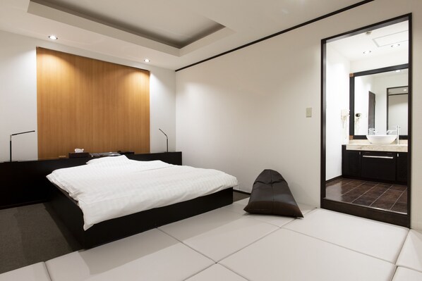 Una habitación de hotel moderna con una cama grande, un cabecero de madera, una pequeña zona de estar y un baño visible a través de una puerta abierta.