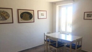 Una stanza con un tavolo bianco e quattro sedie, una finestra con persiane e tre quadri appesi al muro.