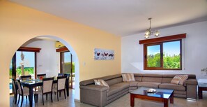 A spacious living area with a large sofa, dining table, and chairs, a ceiling light, and a window with a view of greenery. - Family Villa Short walk to Coral Bay (5 min) Quiet area Large Pool Free bedm A/C (Paphos)