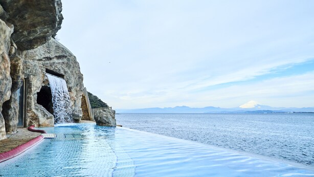 A pool with a waterfall feature, a rocky cliff, and a view of the sea. - Enoshima Hotel (Fujisawa)
