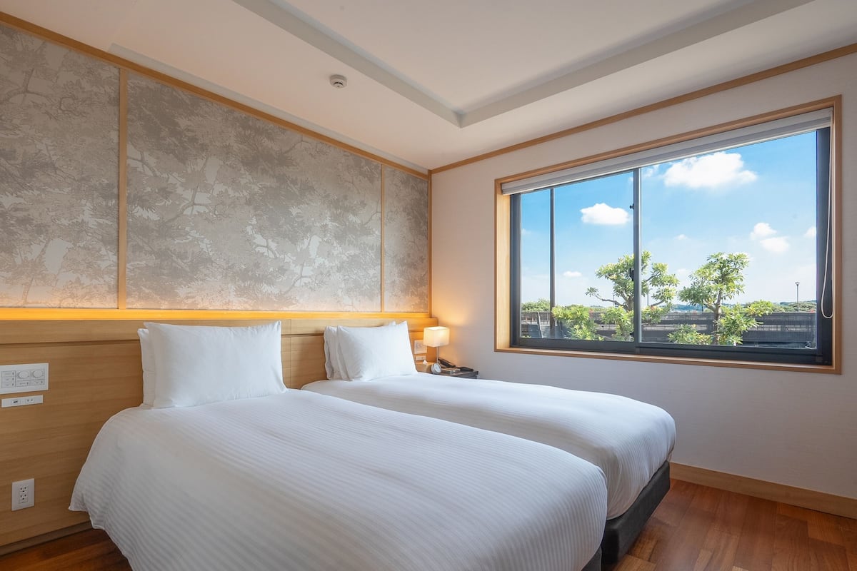 A hotel room with two beds, a large window with a view, a wooden headboard, and a wall-mounted control panel.