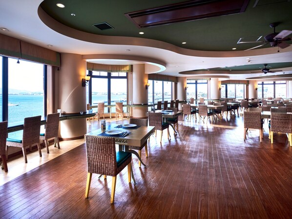 A spacious dining area with high tables and wicker chairs, large windows offering a view of the sea, and a modern ceiling design.
