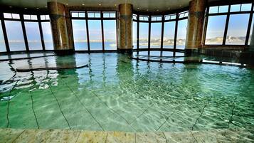 A large indoor swimming pool with clear water, surrounded by large windows offering views of the city skyline.