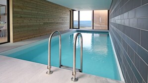An indoor swimming pool with metal railings, wooden walls, and a view of the sea through large windows.