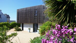 A modern building with a slatted facade, surrounded by lush greenery and flowering plants.
