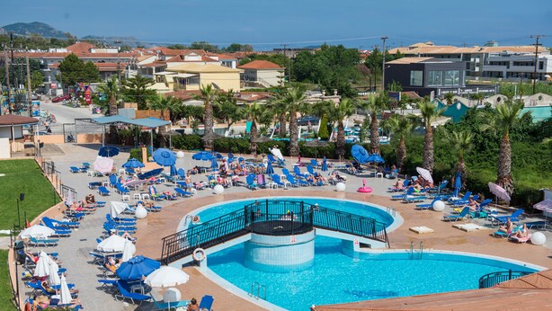 A large outdoor swimming pool with multiple sun loungers and umbrellas, surrounded by palm trees and buildings. - Hotel Alexander the Great (Zakynthos)