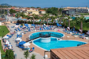 A large outdoor swimming pool with multiple sun loungers and umbrellas, surrounded by palm trees and buildings. - Hotel Alexander the Great (Zakynthos)