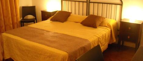 Una cama bien hecha con cabecera, una silla, una mesita de noche y una lámpara.