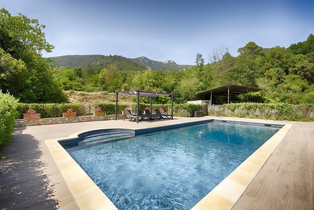 A rectangular swimming pool with a tiled edge, surrounded by a wooden deck. There are lounge chairs and a covered seating area with a pergola. The pool is set against a backdrop of lush greenery and hills.