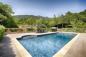 A rectangular swimming pool with a tiled edge, surrounded by a wooden deck. There are lounge chairs and a covered seating area with a pergola. The pool is set against a backdrop of lush greenery and hills. - Mountain View | Stylish Apt | Luxury Bath | Forest (Sales De Llierca)