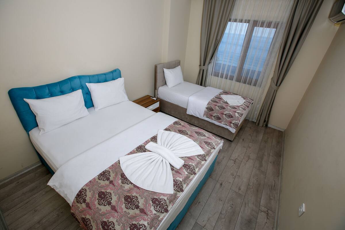 A hotel room with two beds, one with a blue headboard and the other with a grey headboard. Both beds have white linens and decorative towels. The room has wooden flooring and a window with curtains.