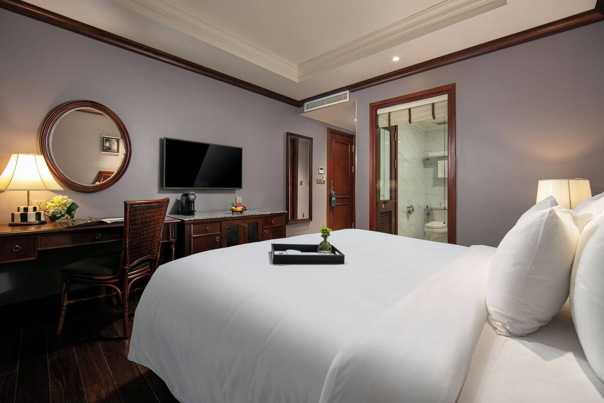 Habitación de hotel con una cama grande, un escritorio con silla, televisor y baño visible a través de una puerta abierta.