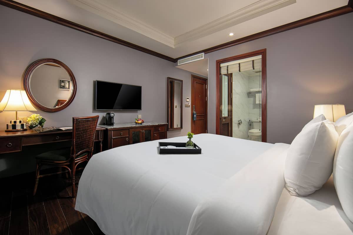 Quarto de hotel com uma cama grande, uma escrivaninha com cadeira, televisão e casa de banho visível através de uma porta aberta.