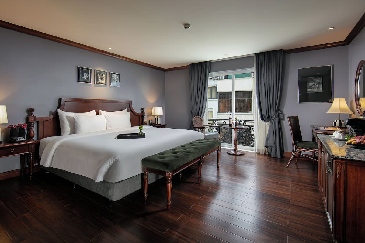 Una habitación de hotel con una cama grande, mesitas de noche, un banco, un escritorio con silla y vistas al exterior.