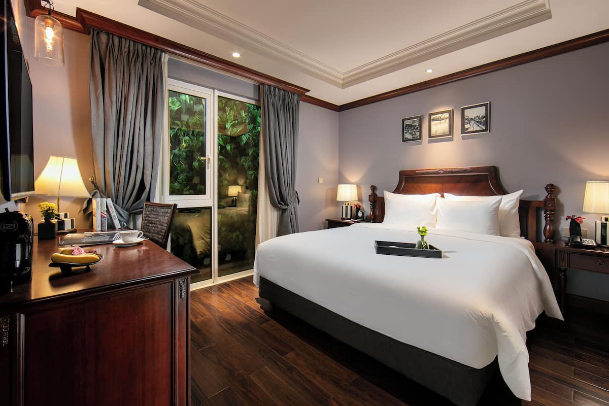 Una habitación de hotel con una cama grande, un escritorio con lámpara, una silla, una mesita de noche con lámpara y una ventana con cortinas.