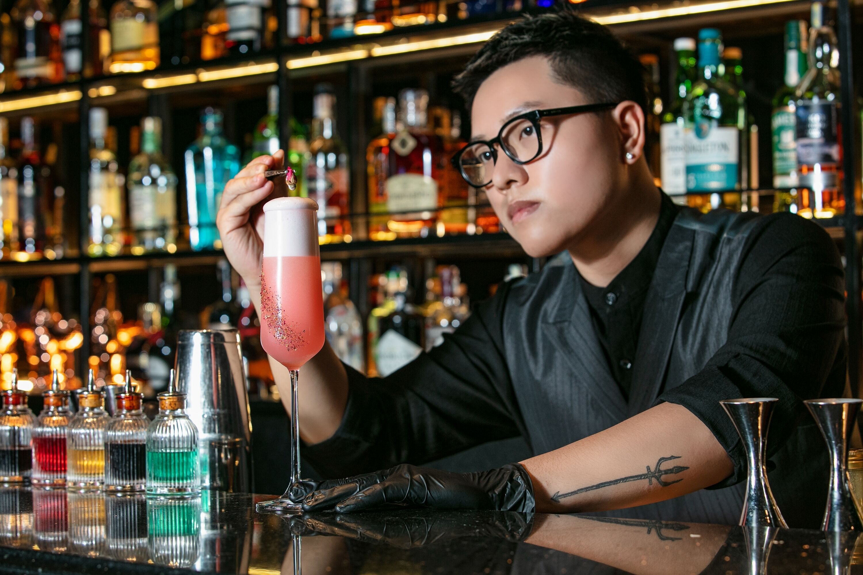 A bartender holding a cocktail with a pink and red color scheme, surrounded by various bottles and bar tools.