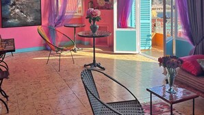 Una habitació amb parets roses, un gran finestral amb cortines morades, una cadira colorida, una taula petita amb un ram de flors i vista a un edifici blau a l'exterior.