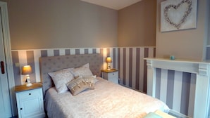 A bedroom with a bed, two bedside tables, a nightstand, a wall-mounted heart decoration, and a white fireplace mantel. - West Flanders Vacation Home (Comines-Warneton)