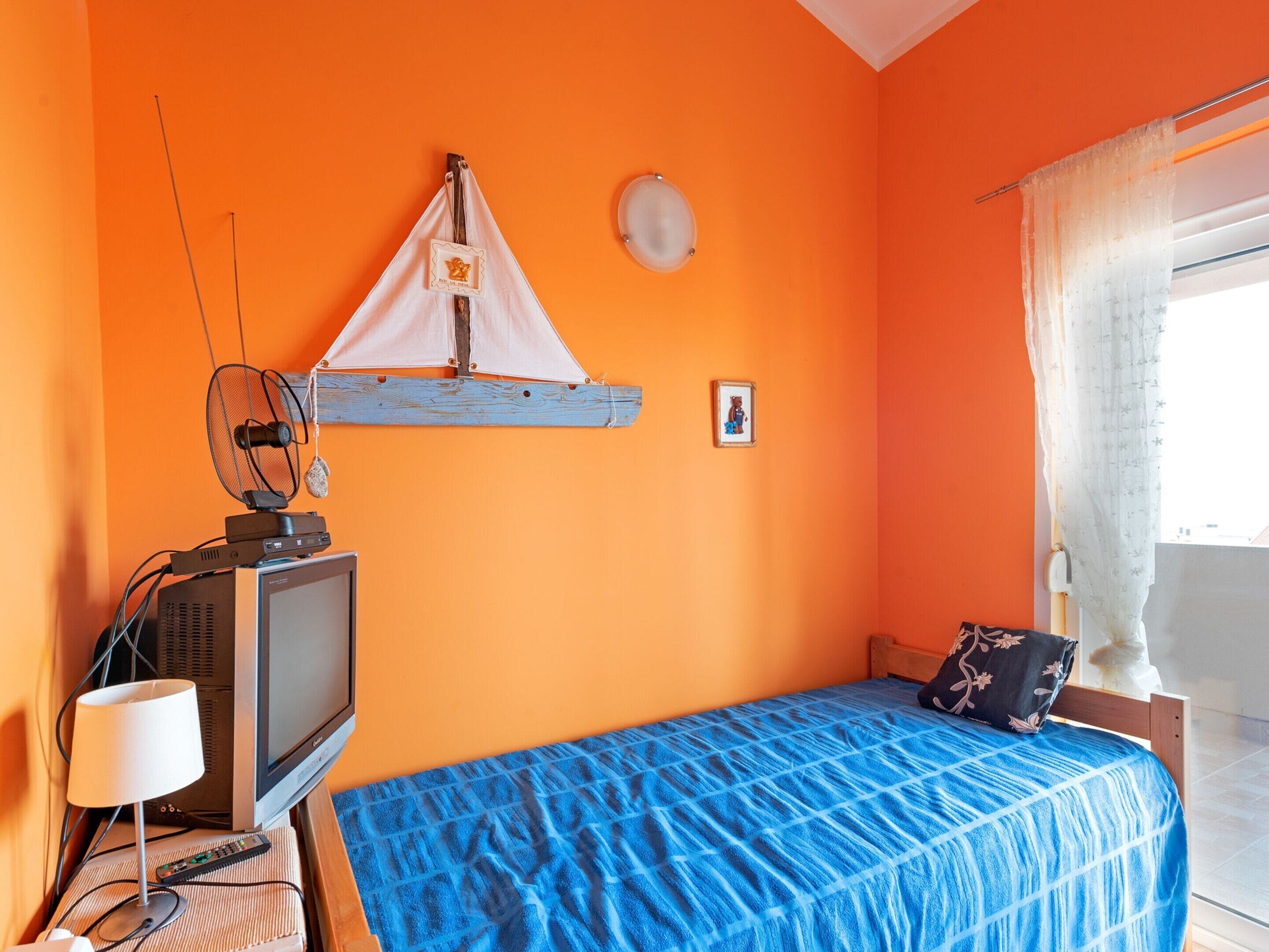 Ein Zimmer mit orangefarbener Wand, einem Bett mit blauer Bettdecke, einem Fernseher auf einem Holzständer, einem kleinen Tisch mit einer Lampe und einem Fenster mit Vorhängen.