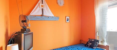 Ein Zimmer mit orangefarbener Wand, einem Bett mit blauer Bettdecke, einem Fernseher auf einem Holzständer, einem kleinen Tisch mit einer Lampe und einem Fenster mit Vorhängen.