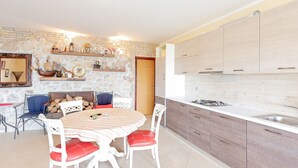 A modern kitchen with white cabinets, a stone wall, and a dining area with a striped tablecloth and red chairs. - Modern Holiday Home in Mandre With Barbeque (Zmijavci)
