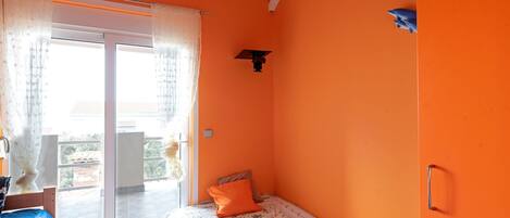 A bedroom with an orange wall, a bed with a patterned bedspread, a small table with a lamp, and a sliding glass door leading to an outdoor area.