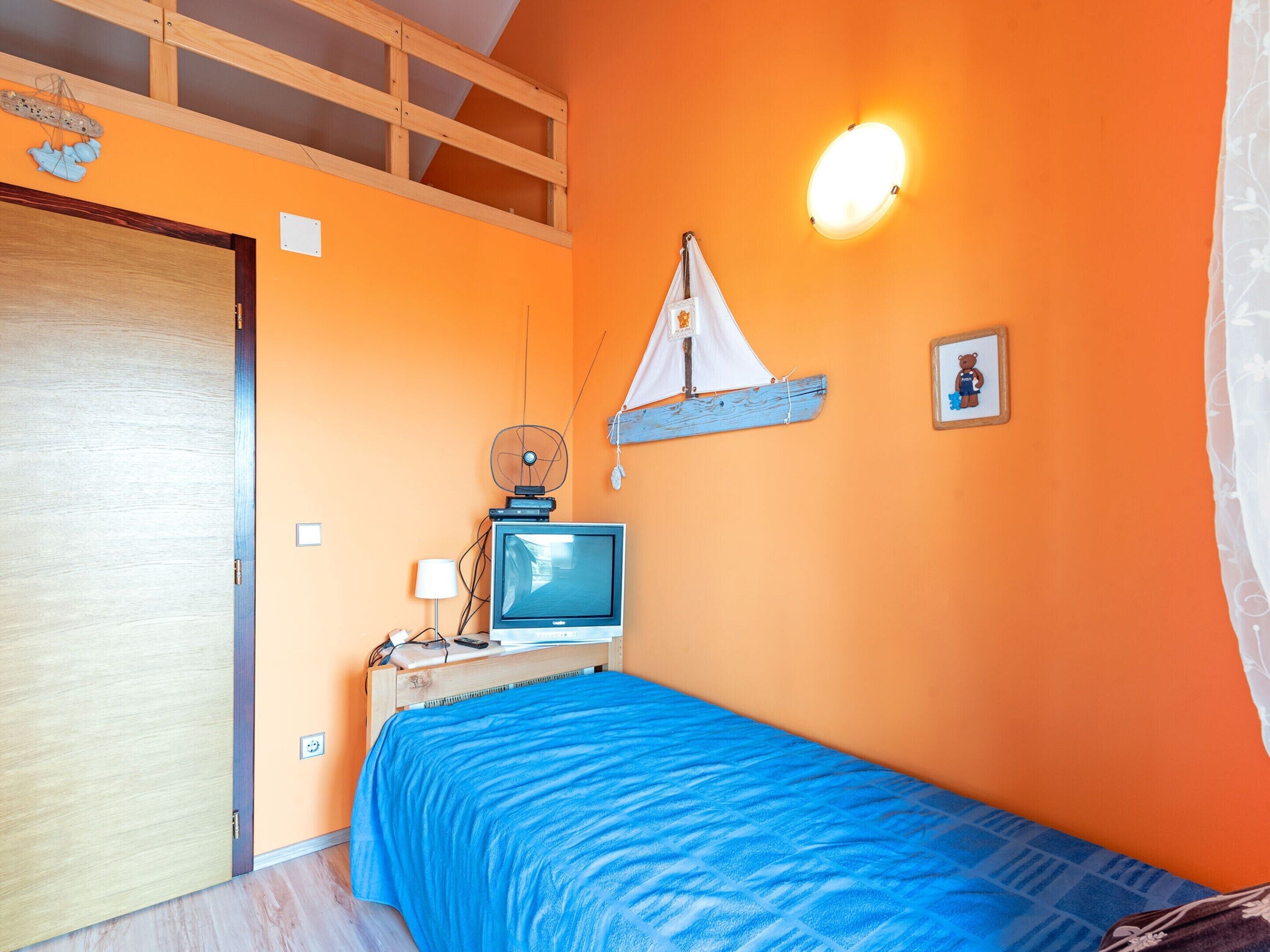 A room with an orange wall, a bed with a blue bedspread, a wooden headboard, a television on a wooden stand, a small sailboat decoration, and a wall-mounted light fixture.