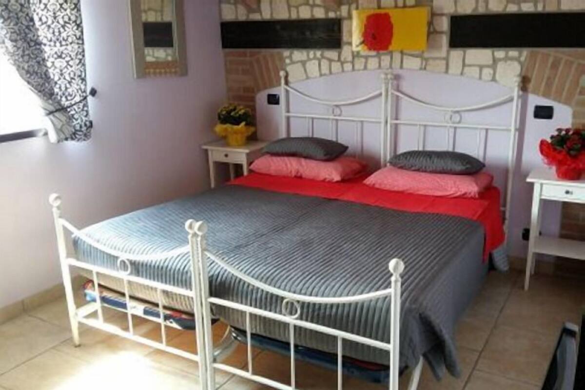 Un dormitorio con estructura de cama de metal blanco, colcha roja y gris, y pared de piedra con un cuadro de colores amarillo y rojo.