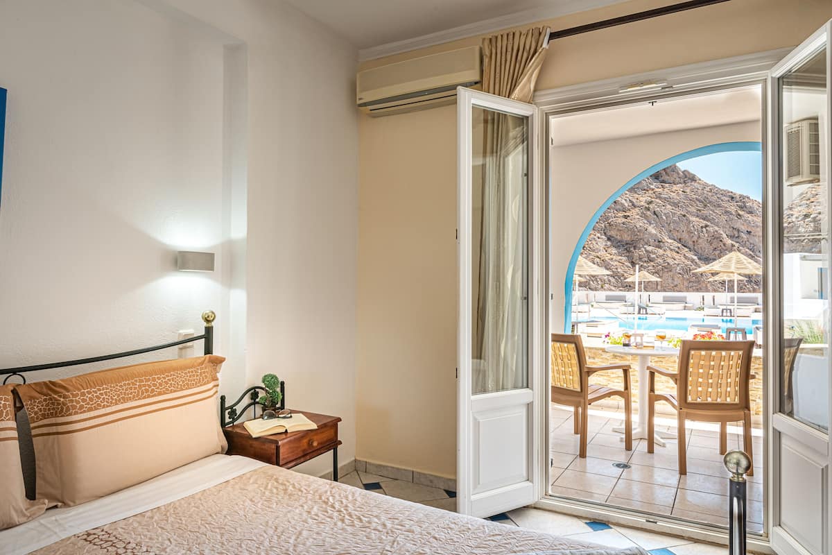 A hotel room with a bed, a nightstand, and an open door leading to a balcony with a pool and chairs.