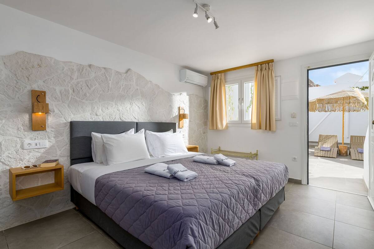 A modern bedroom with a stone accent wall, a large bed with a purple quilt, and a view of the outdoor seating area through a glass door.