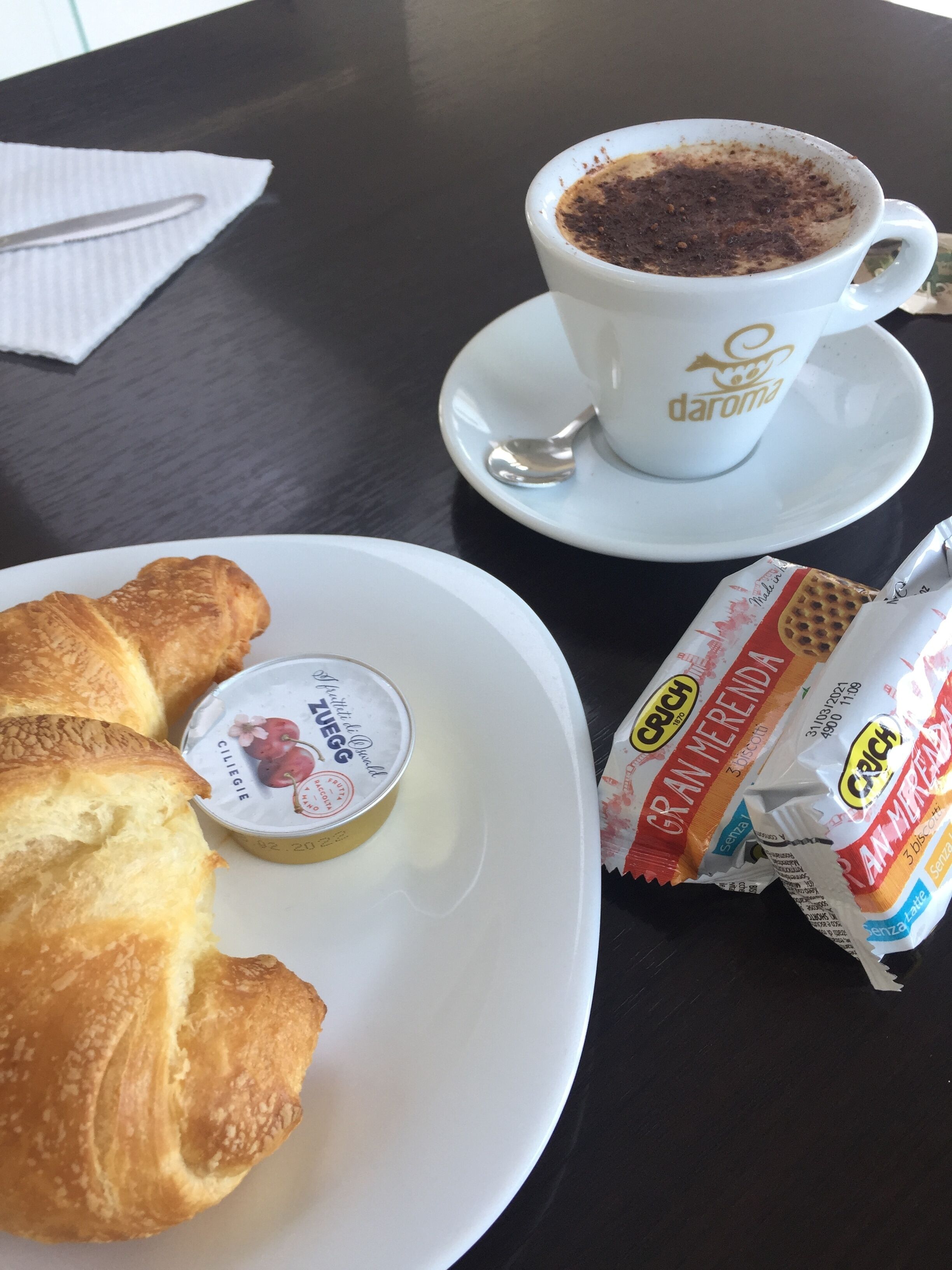 Un tavolo con un croissant, una tazza di caffè, un piccolo vasetto di marmellata e un pacchetto di biscotti.