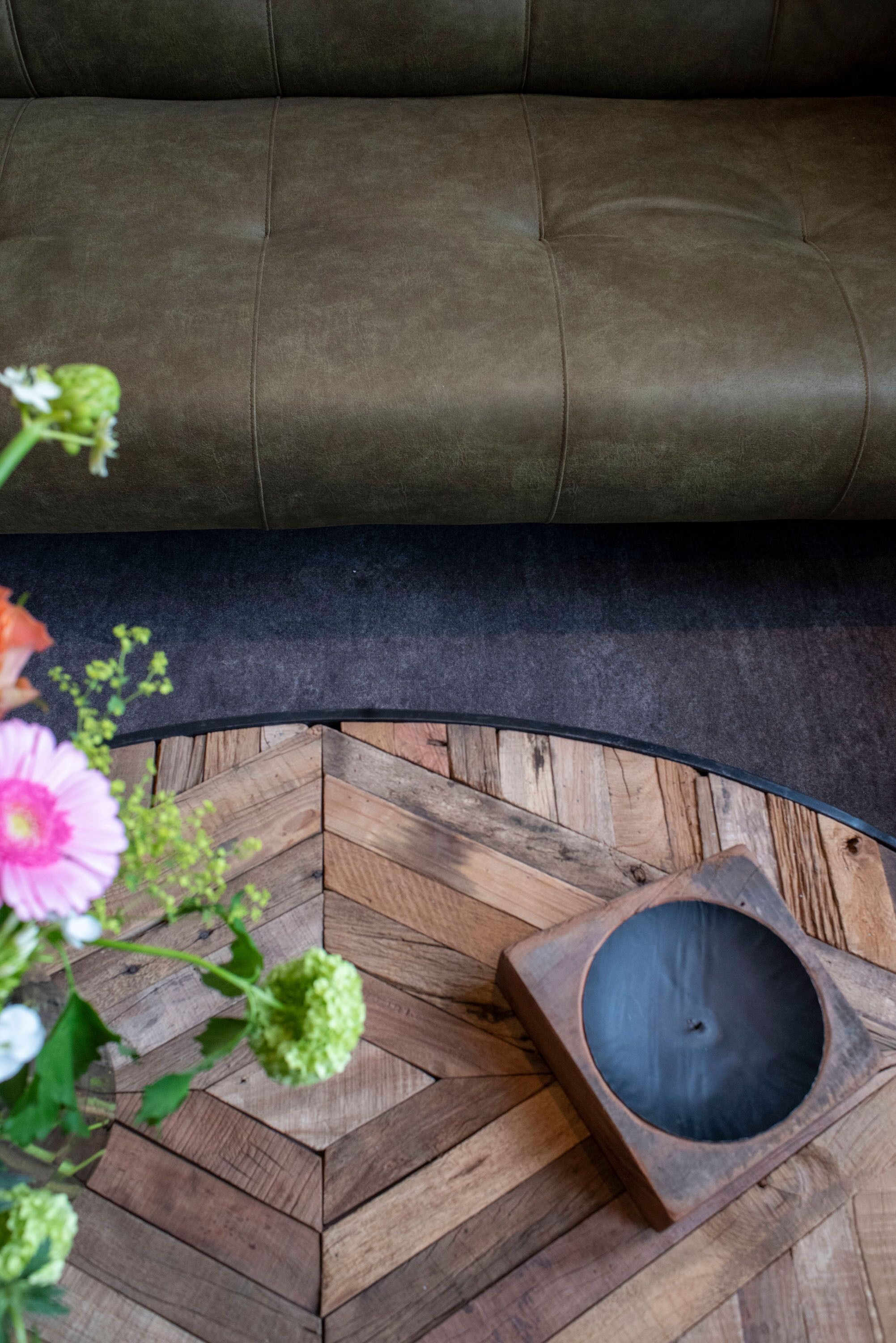 A brown leather sofa, a wooden herringbone floor, and a small wooden table with a black circular object on it.
