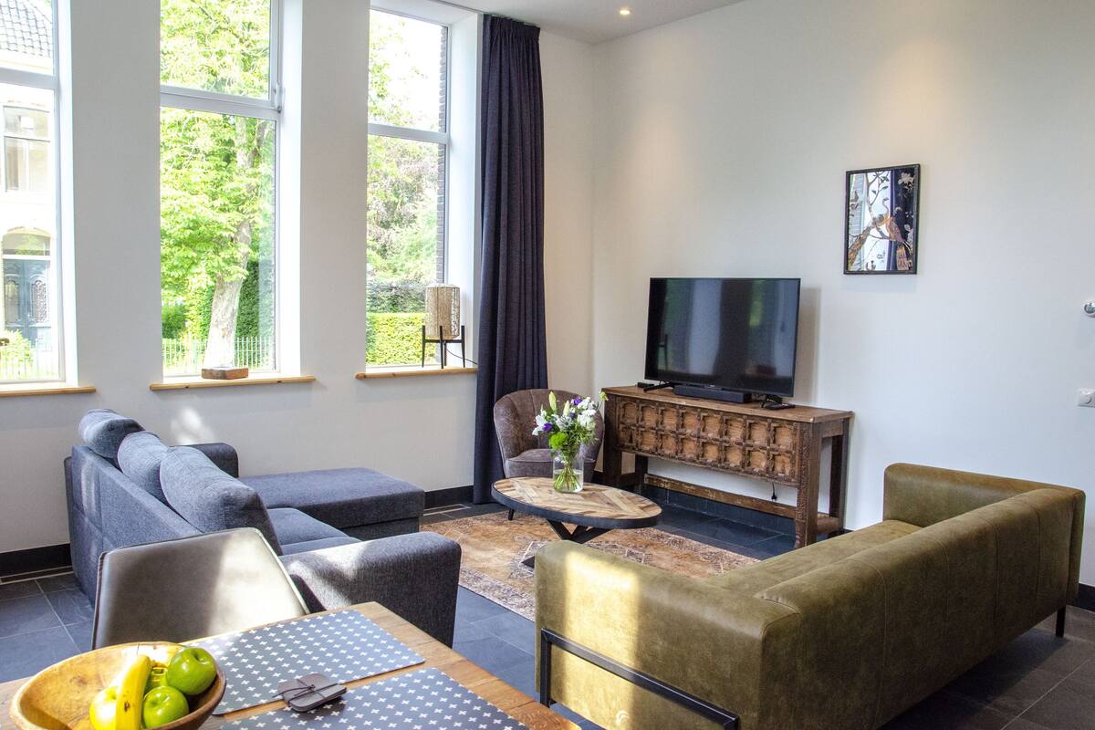 Ein modernes Wohnzimmer mit einer Couch, einem Sessel und einem Couchtisch mit Blumen. Ein Flachbildfernseher ist an der Wand befestigt, und es gibt ein Fenster mit Blick auf Grünflächen.