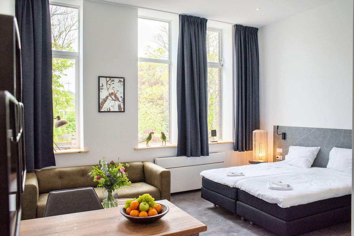 Ein Hotelzimmer mit einem Bett, einem Sofa, einem Esstisch mit Obst und großen Fenstern mit Vorhängen.