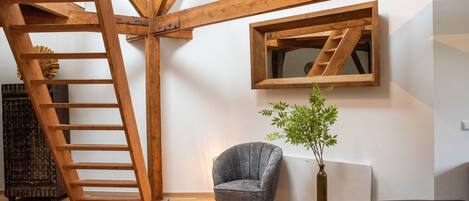 A room with a wooden ladder, a grey armchair, a potted plant, and a black ottoman.