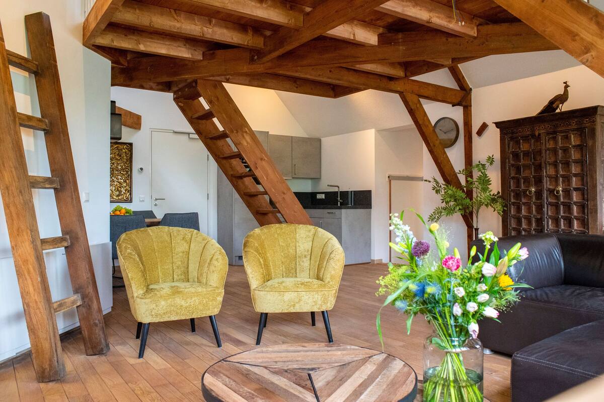 Ein Loft-Wohnbereich mit freiliegenden Holzbalken, einer Treppe, zwei Sesseln, einem Couchtisch, einer Blumenvase und einem schwarzen Ledersofa.