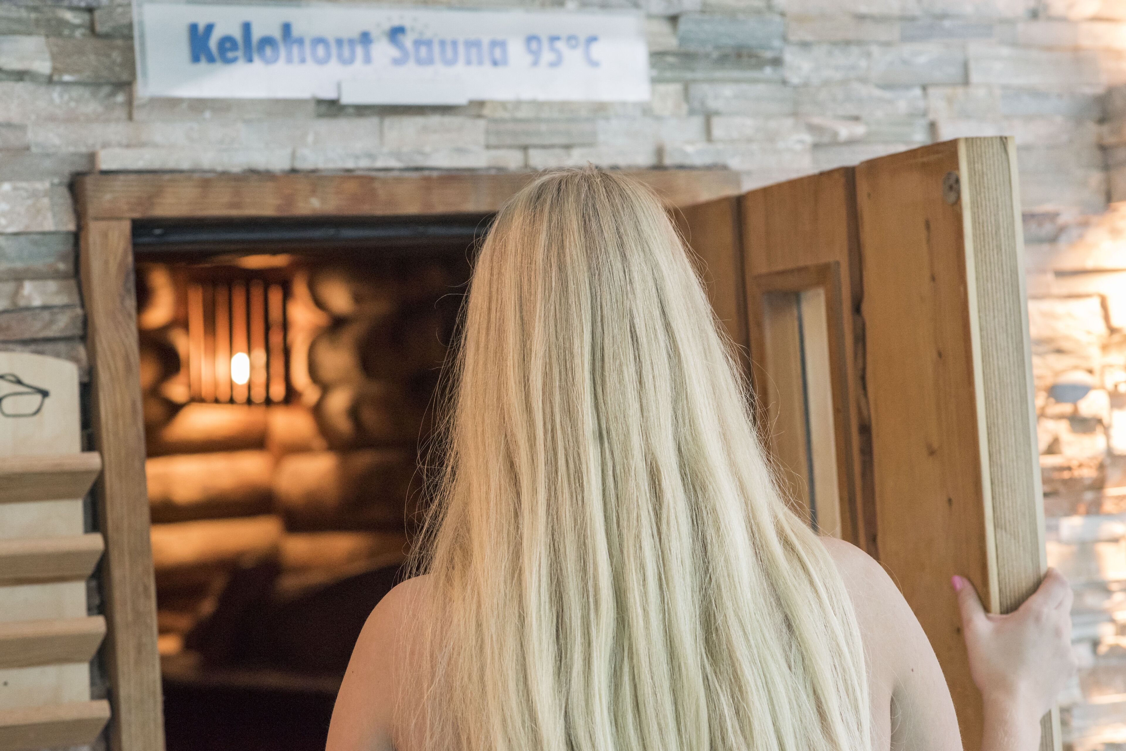 A person with long blonde hair opening a wooden door to a room with a stone wall and a sign indicating a sauna at 95°C.
