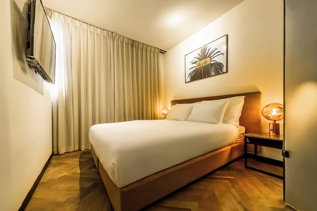 Ein Hotelzimmer mit einem Bett, einem Wand-Fernseher, einer Nachttischlampe und einem gerahmten Bild an der Wand.