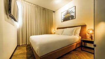 Habitación de hotel con una cama, televisor montado en la pared, lámpara de noche y un cuadro enmarcado.